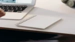 Notion AI — real photo of a blank spiral notebook on a desk beside a laptop