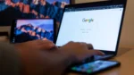 optimize website for ai search — Close-up of hands typing on a laptop showing the Google search homepage on screen, with a second monitor displaying a mountain wallpaper in the background.