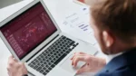free ai search optimization tools — Over-the-shoulder view of a man in a blue suit working on a silver laptop showing an analytics dashboard with candlestick-style charts, printed reports lying on the desk beside him