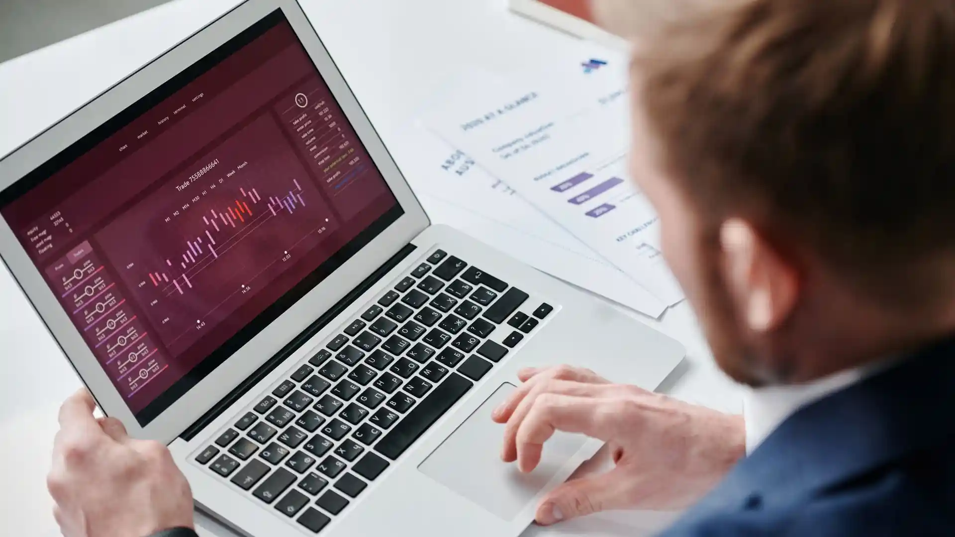 free ai search optimization tools — Over-the-shoulder view of a man in a blue suit working on a silver laptop showing an analytics dashboard with candlestick-style charts, printed reports lying on the desk beside him