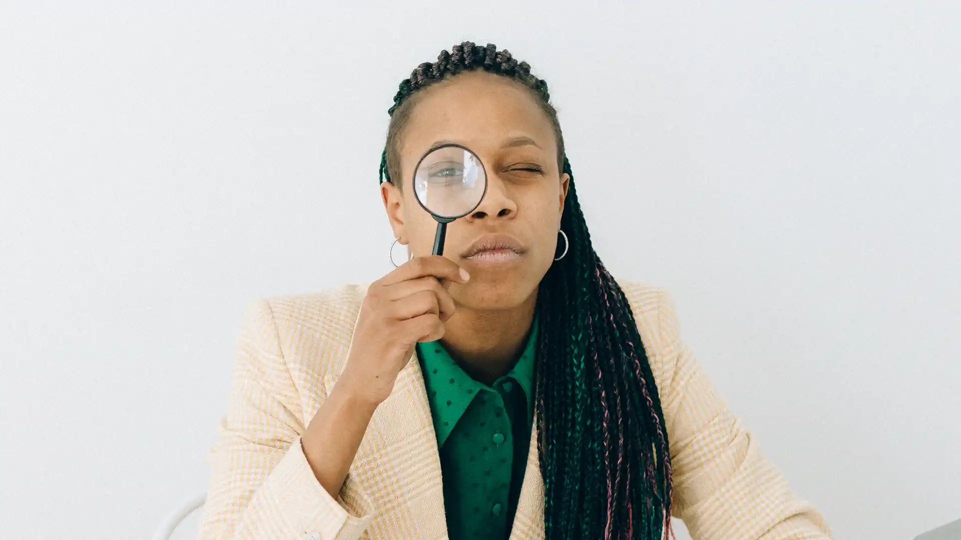 what is search optimization — Woman with long braided hair in a beige blazer and green polka dot blouse holding a magnifying glass over her right eye against a plain light background