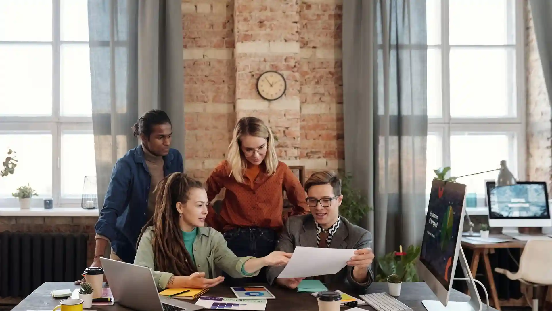 answer engine optimization best practices — Diverse team of four professionals discussing work around a desk in a modern brick-walled office, looking at a document, laptop and monitor