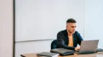 ai ad copy generator — a focused businessman typing on a laptop at an office desk in front of a whiteboard.