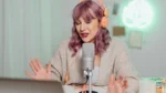 free ai noise remover — a woman with purple hair wearing orange headphones speaks into a studio microphone at her desk.