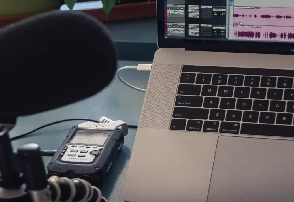 AI podcast transcript generator — real photo of a podcast recording setup with a microphone, Zoom H4n Pro recorder, and a MacBook Pro showing audio editing waveforms