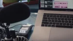 AI podcast transcript generator — real photo of a podcast recording setup with a microphone, Zoom H4n Pro recorder, and a MacBook Pro showing audio editing waveforms