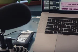 AI podcast transcript generator — real photo of a podcast recording setup with a microphone, Zoom H4n Pro recorder, and a MacBook Pro showing audio editing waveforms