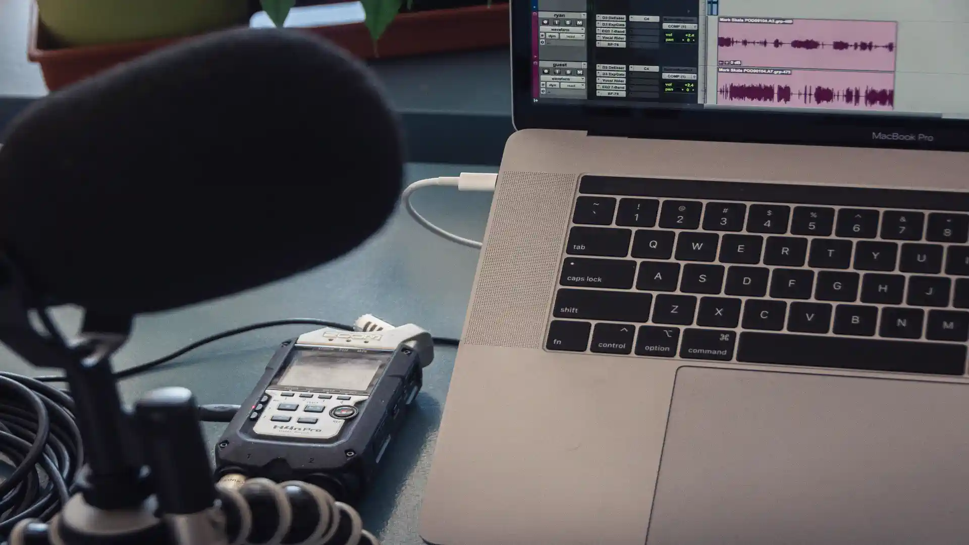 AI podcast transcript generator — real photo of a podcast recording setup with a microphone, Zoom H4n Pro recorder, and a MacBook Pro showing audio editing waveforms