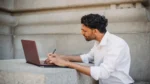 AI email writer free — real photo of a freelancer in a white shirt writing on a laptop outdoors, holding a pen