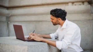 AI email writer free — real photo of a freelancer in a white shirt writing on a laptop outdoors, holding a pen