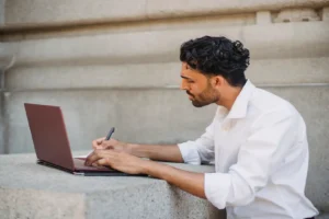 AI email writer free — real photo of a freelancer in a white shirt writing on a laptop outdoors, holding a pen