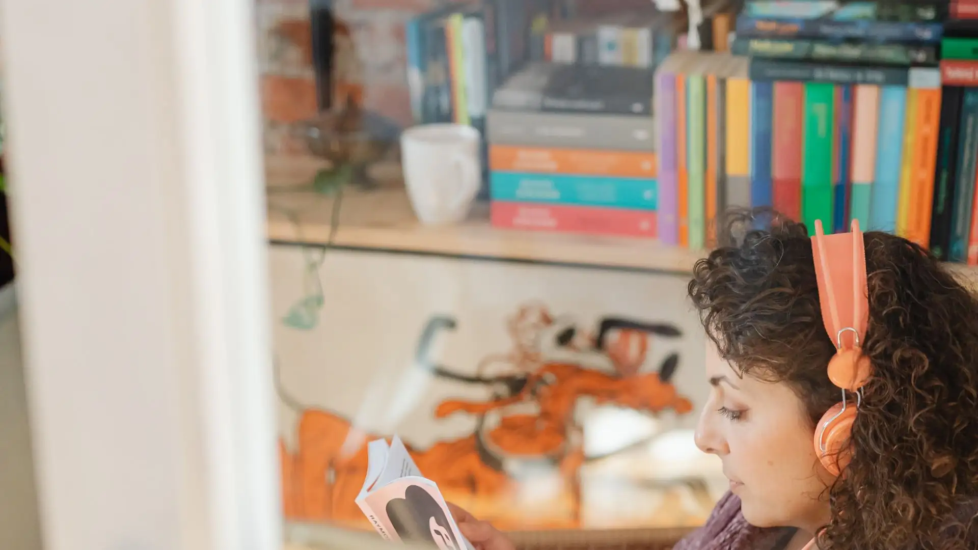 ai audiobook narrator free — Woman with curly hair wearing orange headphones reading a book beside a bookshelf with colorful books and a white mug