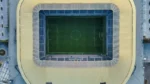 connections sports edition hints — Aerial top-down view of a soccer stadium with a green pitch, white field markings, tiered stands and a pale yellow roof.