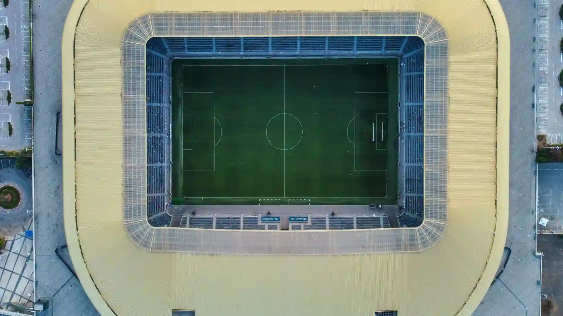 connections sports edition hints — Aerial top-down view of a soccer stadium with a green pitch, white field markings, tiered stands and a pale yellow roof.