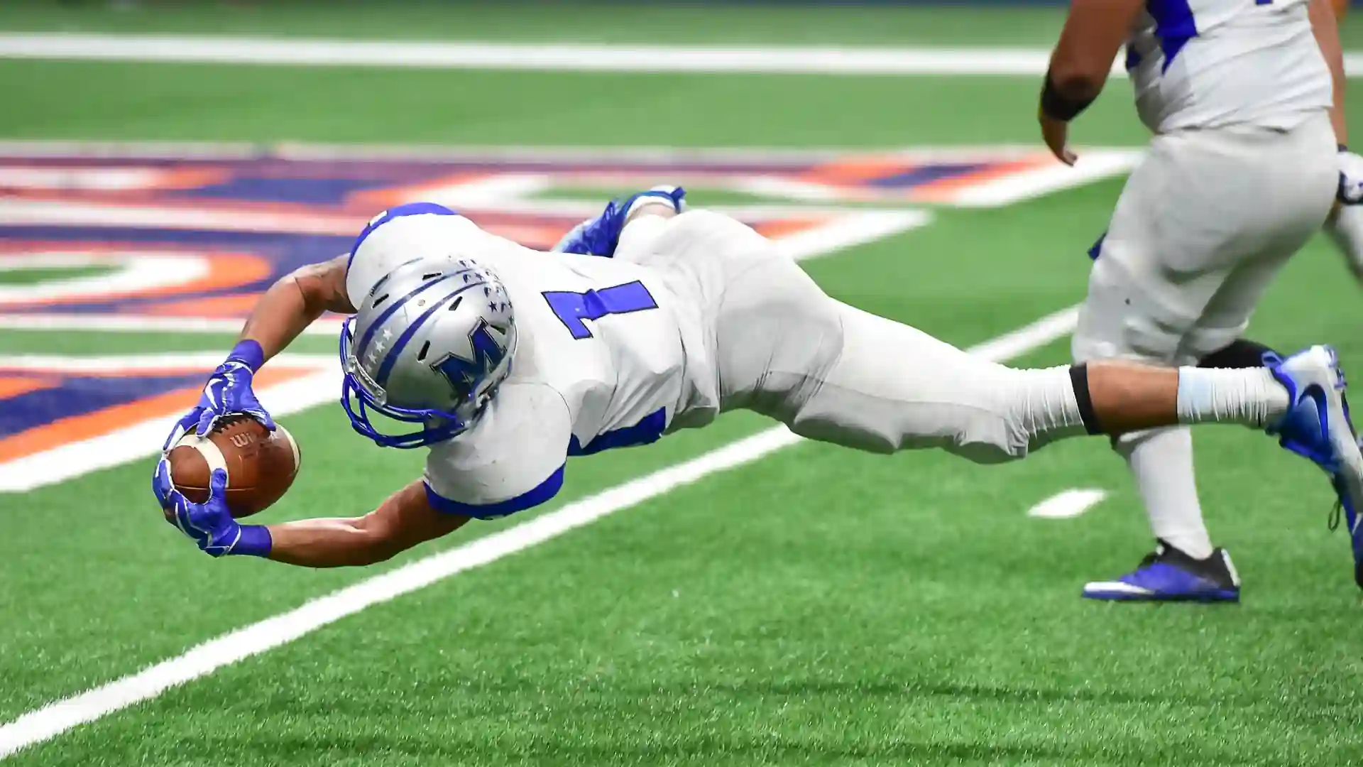 connections sports edition answers today — American football player in a white and blue uniform diving mid-air while holding the football during a game on a green field.