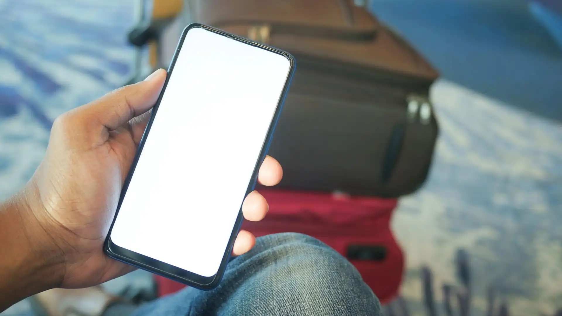 ai voice translator app — Hand holding a smartphone with a blank white screen in an airport, with a red suitcase and travel bag blurred in the background