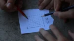 connections game tips and tricks — Hands of two people solving a word search puzzle on a torn sheet of paper with pencils, with found words circled.