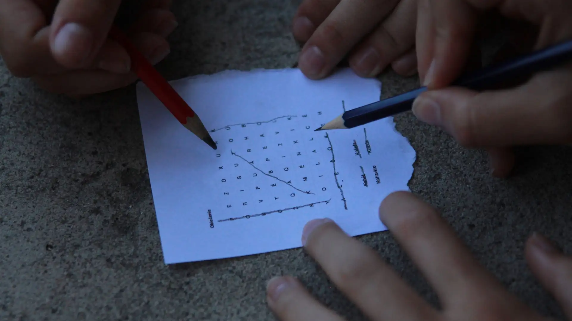 connections game tips and tricks — Hands of two people solving a word search puzzle on a torn sheet of paper with pencils, with found words circled.