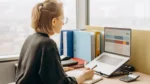 ai spreadsheet formula generator — Woman with glasses working at an office desk with a laptop showing a spreadsheet, a notebook, a calculator and colorful binders