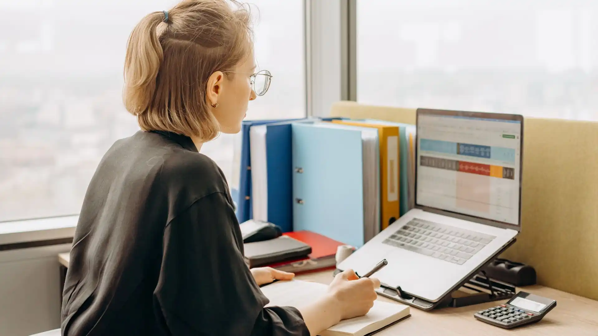 ai spreadsheet formula generator — Woman with glasses working at an office desk with a laptop showing a spreadsheet, a notebook, a calculator and colorful binders