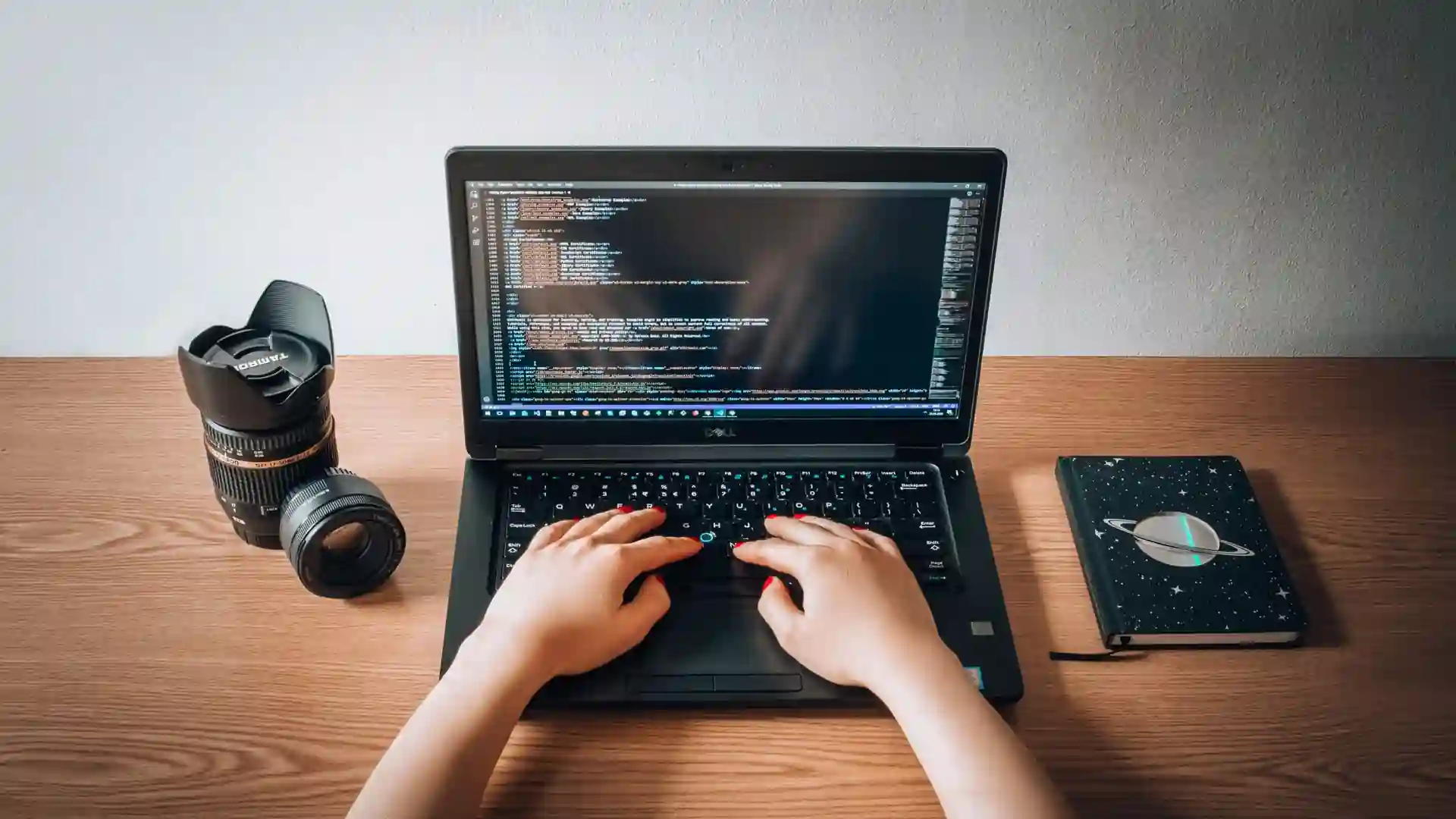 A person's hands typing on a laptop with a code editor open, a camera lens and a notebook on the desk.