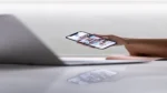 AI social media scheduling tools — real photo of a hand holding a smartphone showing a photo feed beside a laptop